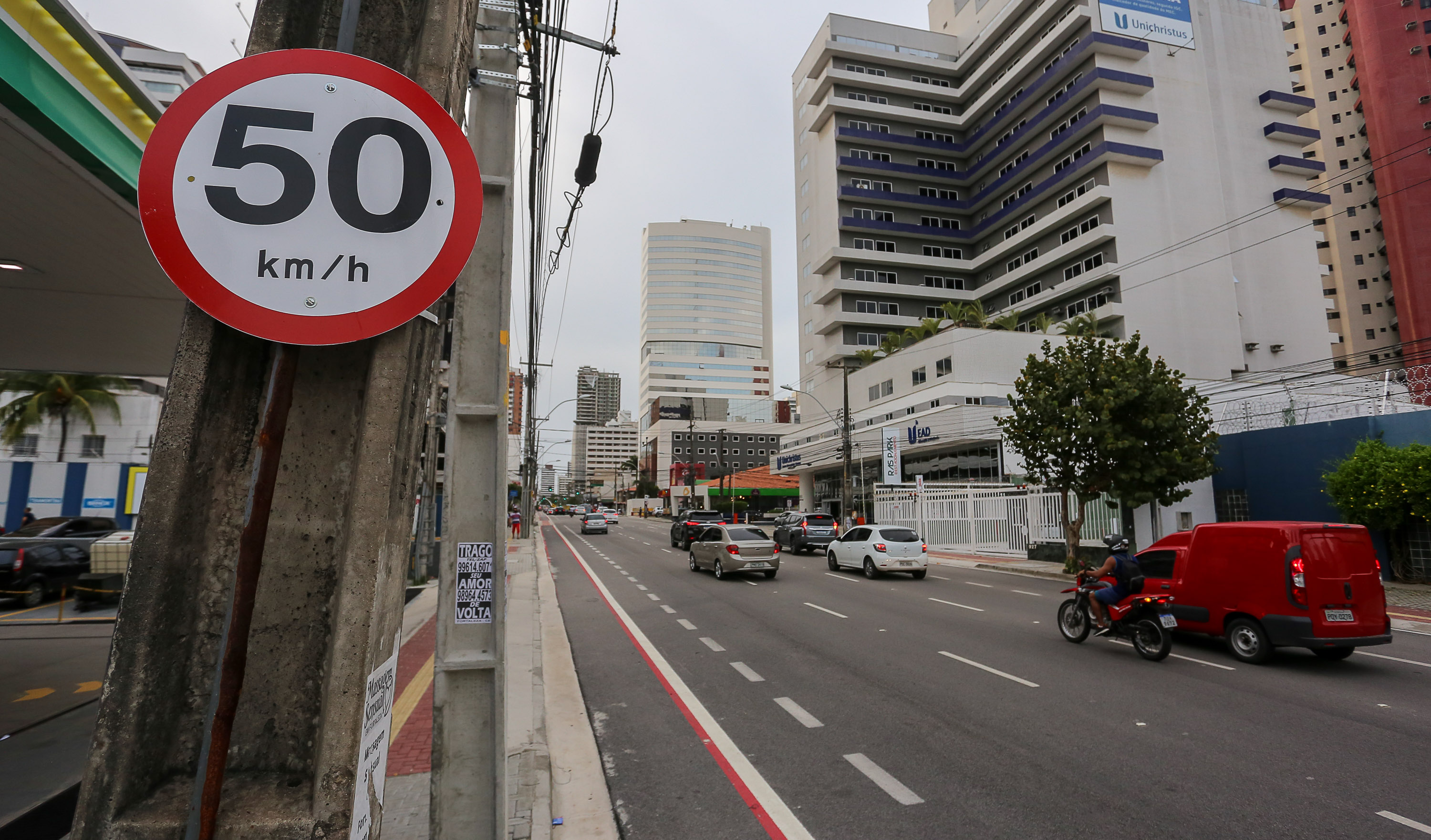 placa de 50 km/h em primeiro plano do lado esquerdo pregada em poste e carros passando em avenida do lado direito